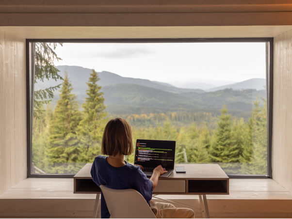 Woman working on a laptop out inside her home with a window showing wilderness Woman working on a laptop out inside her home with a window showing wilderness
